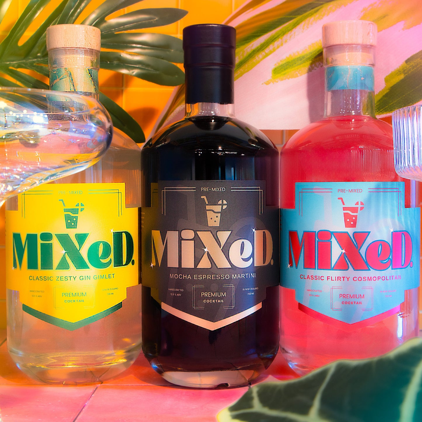 Three bottles of 'Mixed' premixed cocktail drinks on a colourful background with plant leaves, miami vice themed.