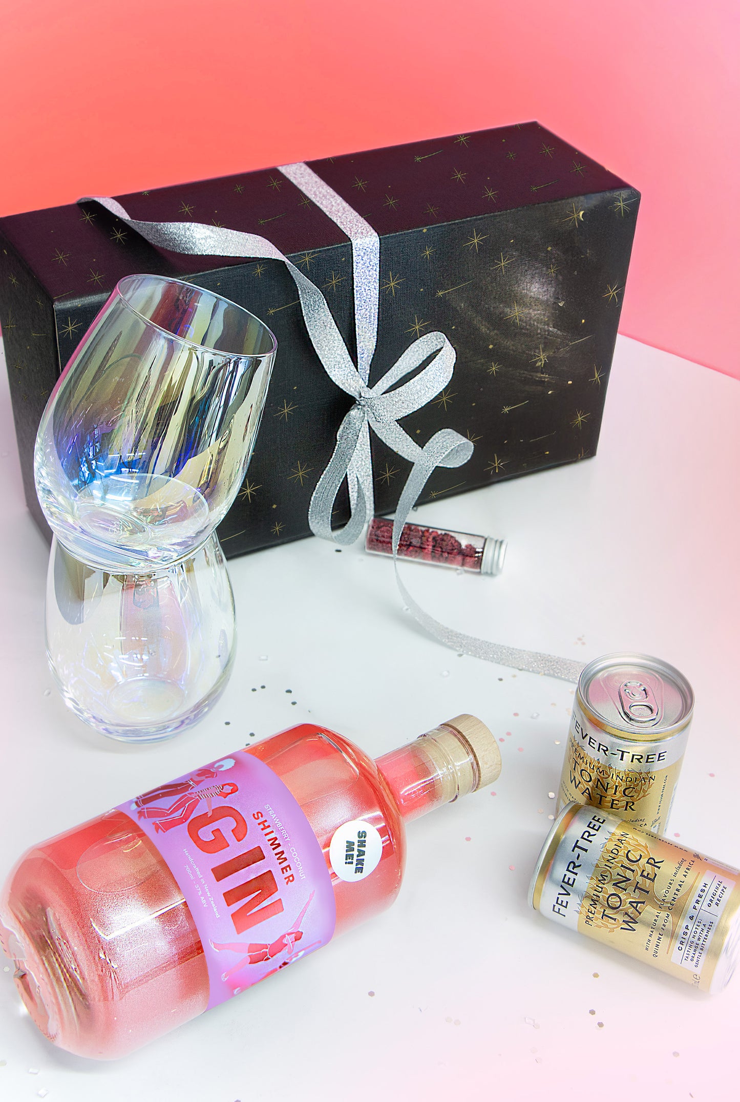 Gin bottle, tonic water can, glass, and gift box on a white surface with pink gradient background