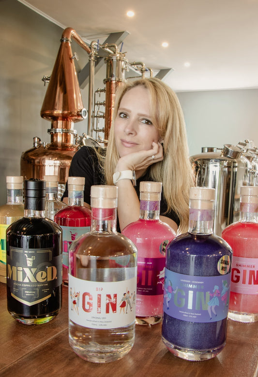 A women distiller sitting behind her gin and cocktail products in a distillery setting.
