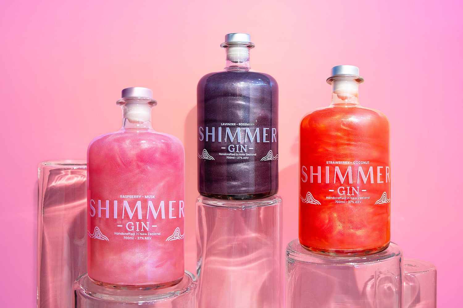 Three bottles of SIP Distilling’s Shimmer Gin in pink and purple hues displayed side by side with fruit garnish, photographed on a bright pastel background.