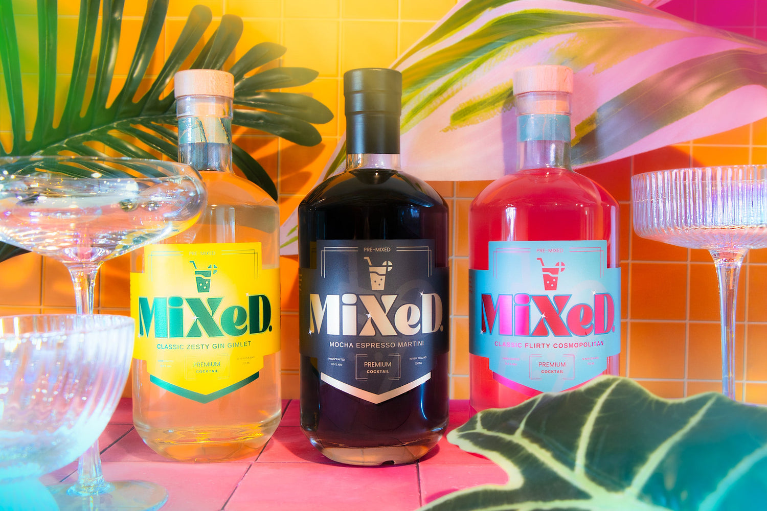 Three MIXED premixed cocktails displayed with vibrant tropical leaves and glassware against a bright colourful backdrop.