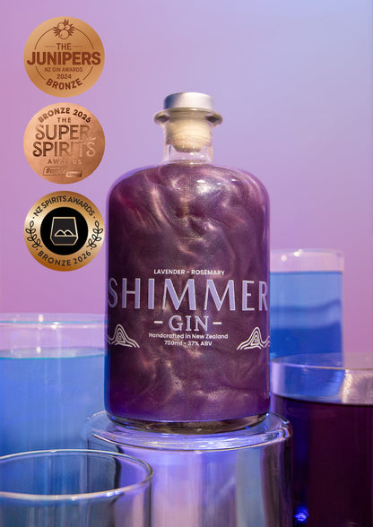 A bottle of Shimmer Gin lavender-rosemary flavored liquefied gin, shown on a metallic pedestal with blue and purple cocktails in the background and award badges on the left.