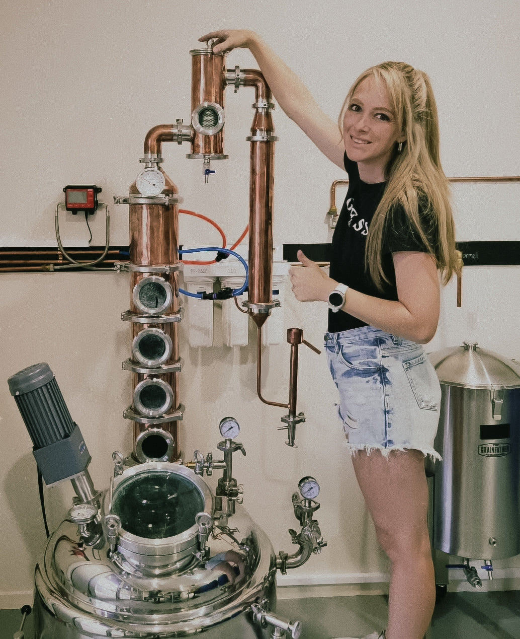 Person standing next to a copper still in a distillery setting