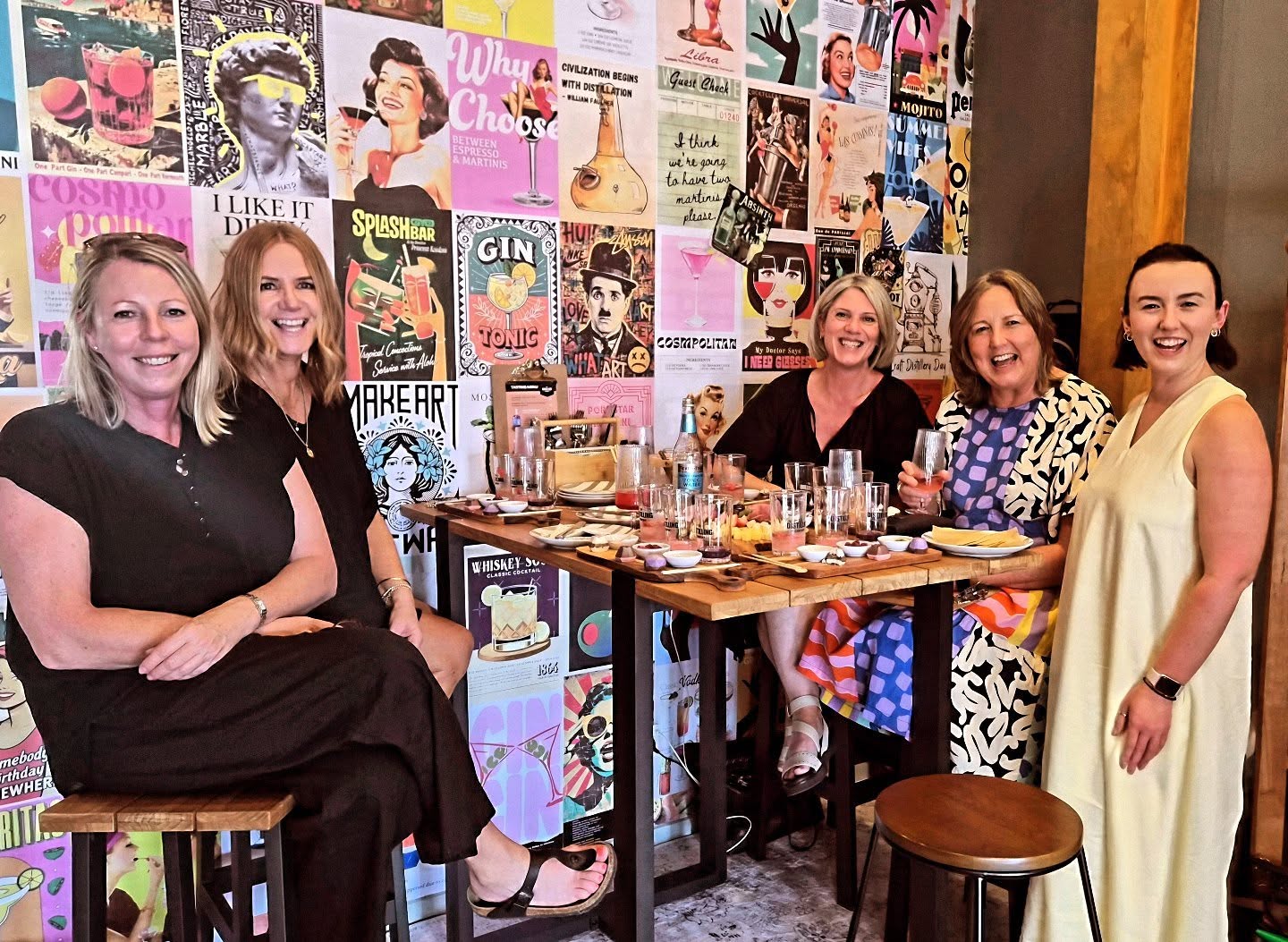 Group of women sitting around a table in a room with a colorful wall of posters.
