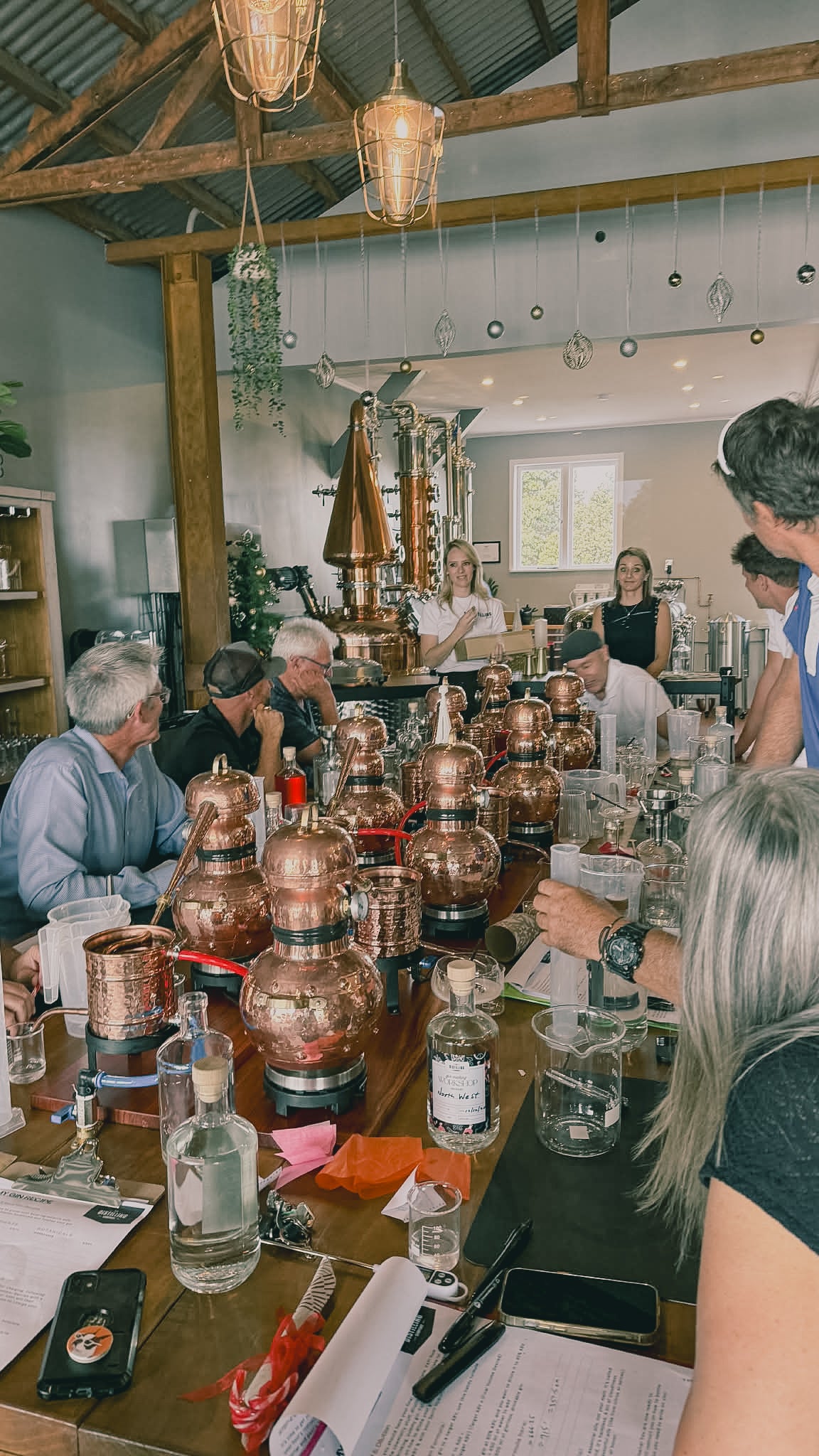 Group of people around a table with copper stills and bottles, likely in a distillery setting.