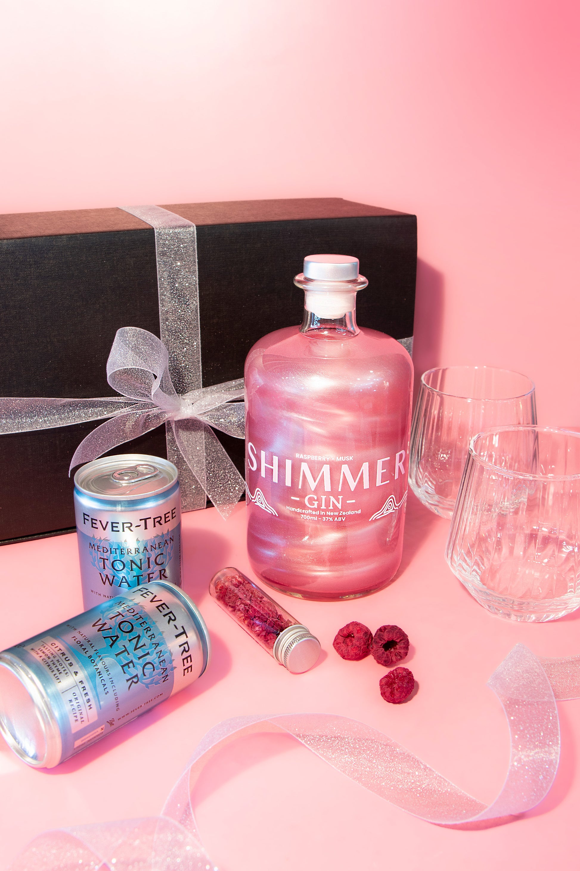 Shimmer gin bottle with tonic water can, raspberries, and glasses on a pink background