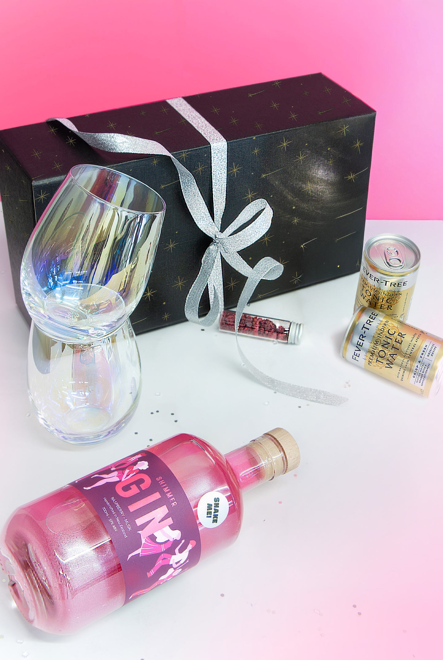 Bottle of pink gin with a glass, gift box, and can on a white surface with pink gradient background