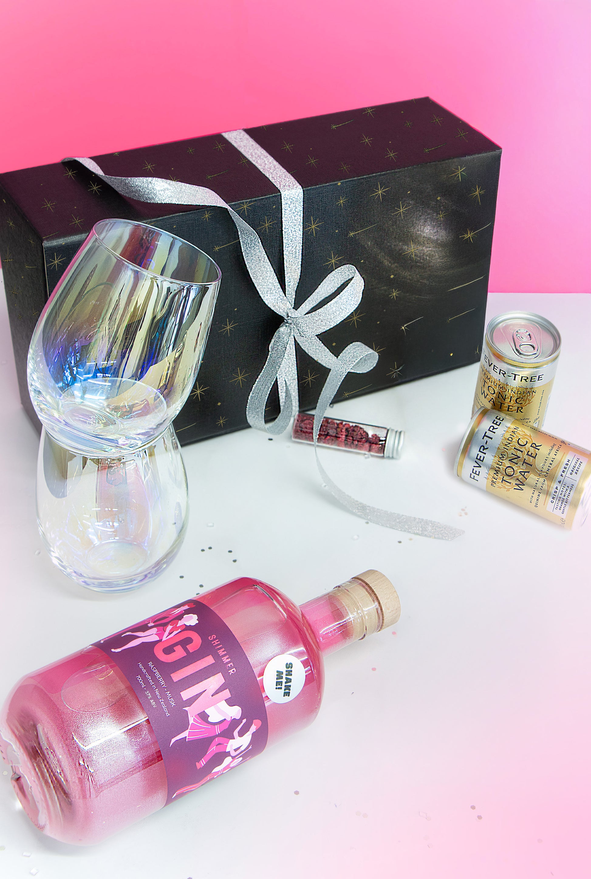 Bottle of pink gin with a glass, gift box, and can on a white surface with pink gradient background