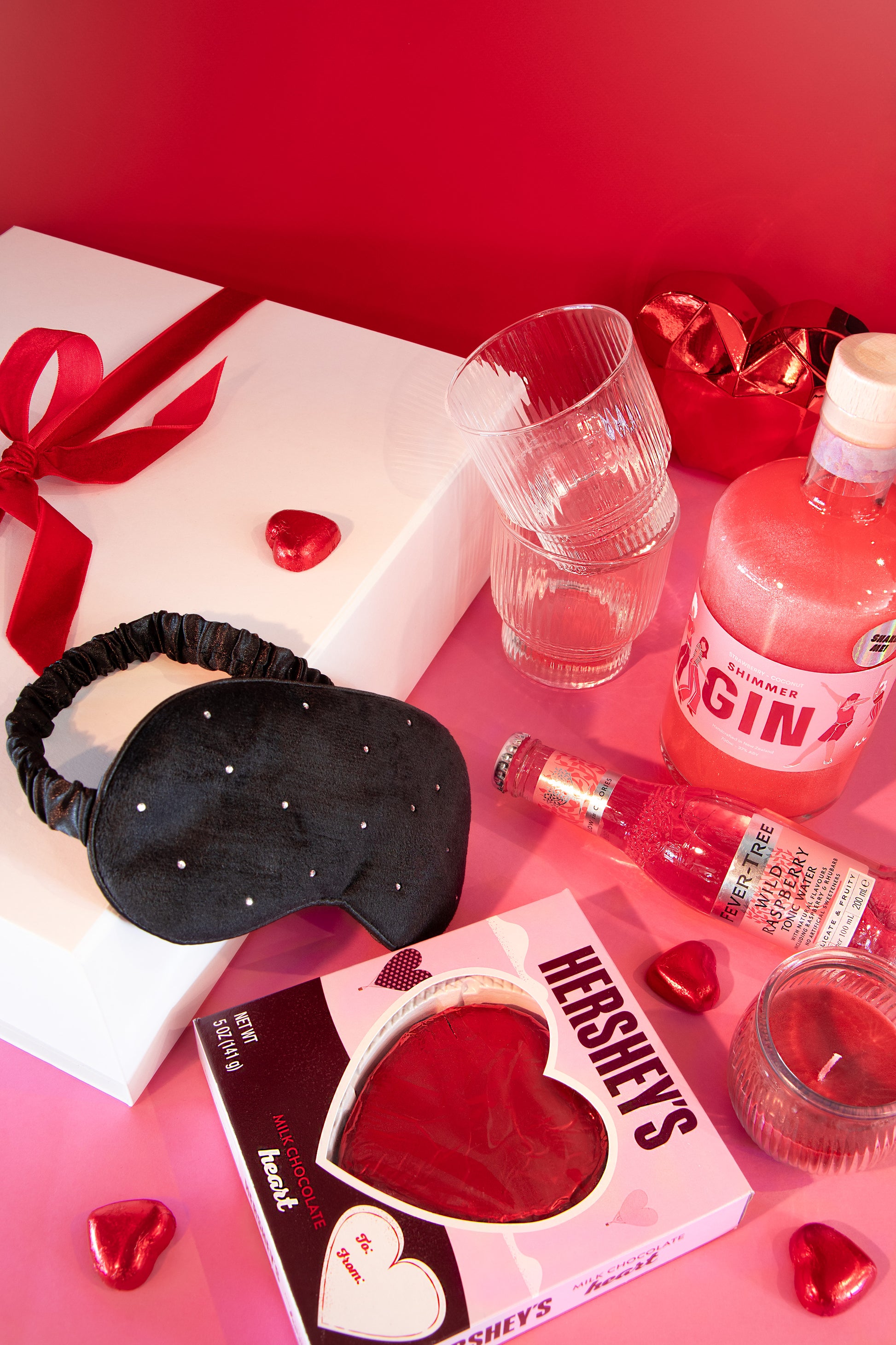 Valentine's Day-themed items including a sleep mask, Hershey's chocolate, and a bottle of gin on a pink surface.