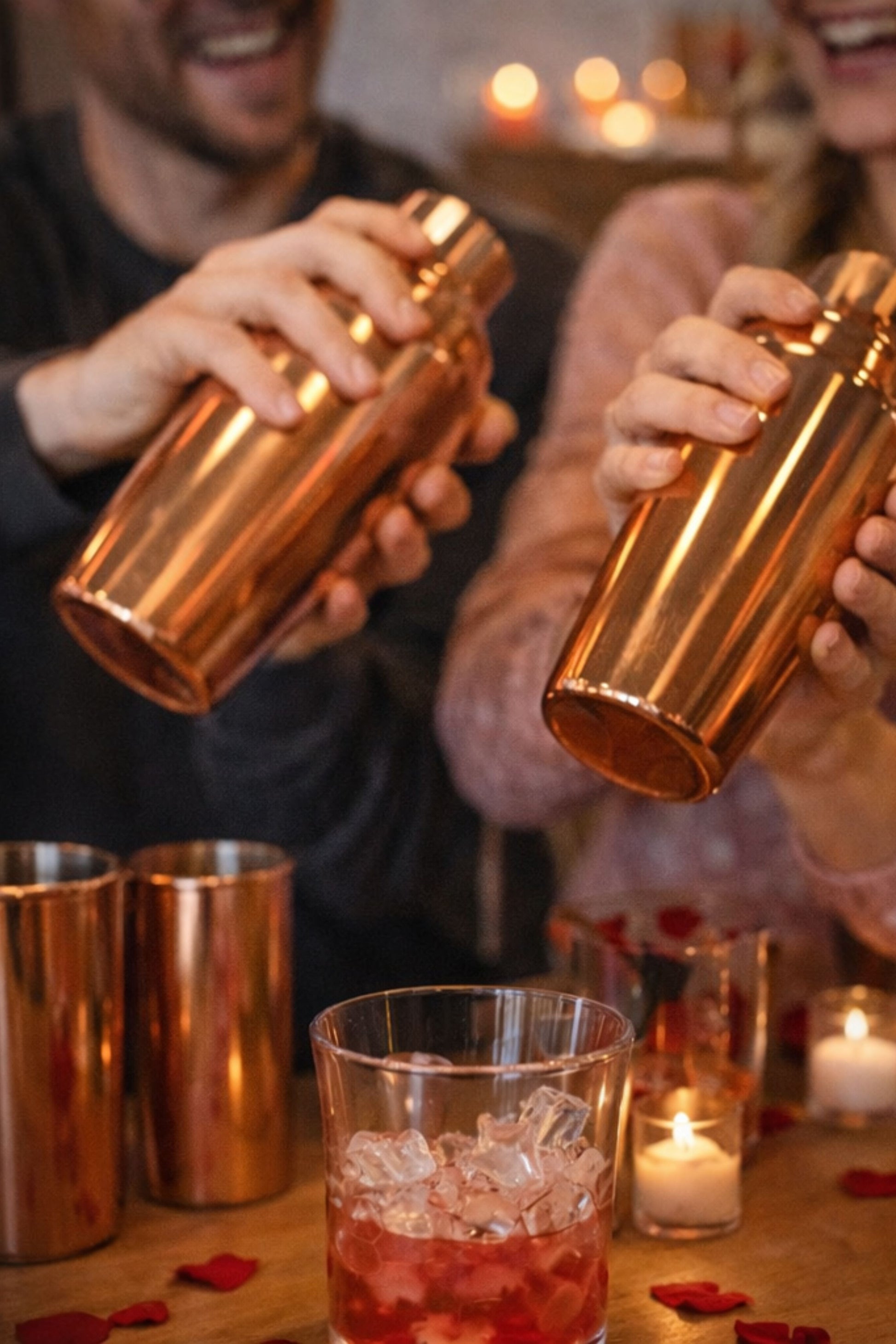 Two people pouring drinks from copper shakers into glasses with a warm, candlelit background.